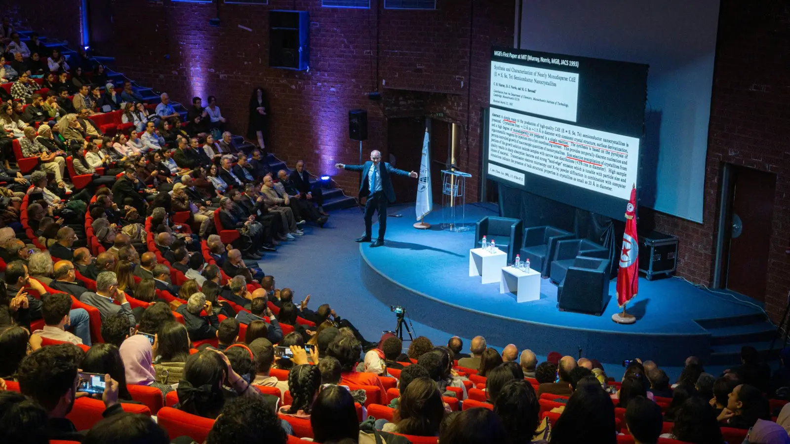 A speaker on a blue-lit stage beside a large screen, facing a full tiered auditorium