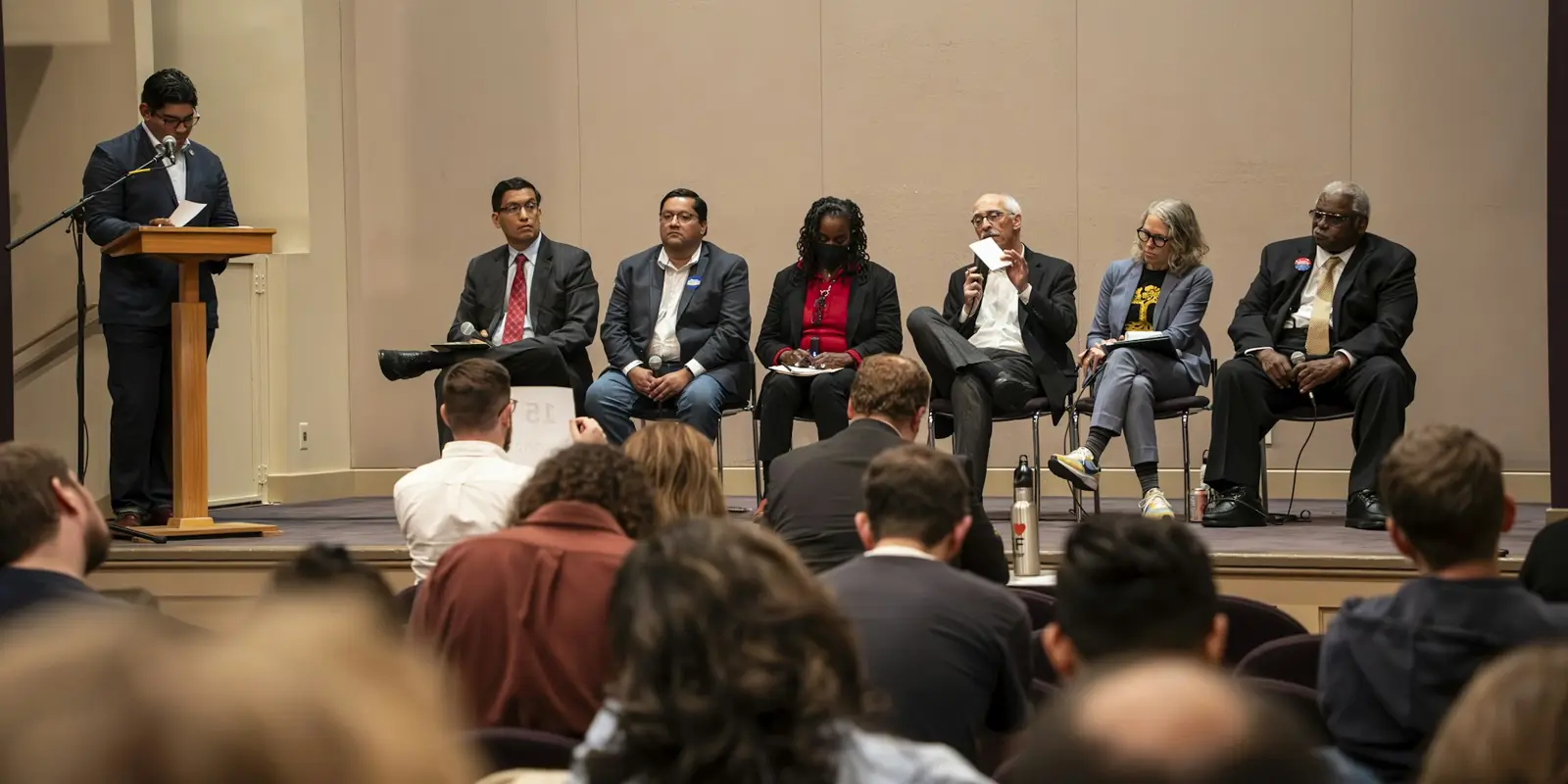 A panel of speakers seated on stage beside a lectern, with the audience in the foreground